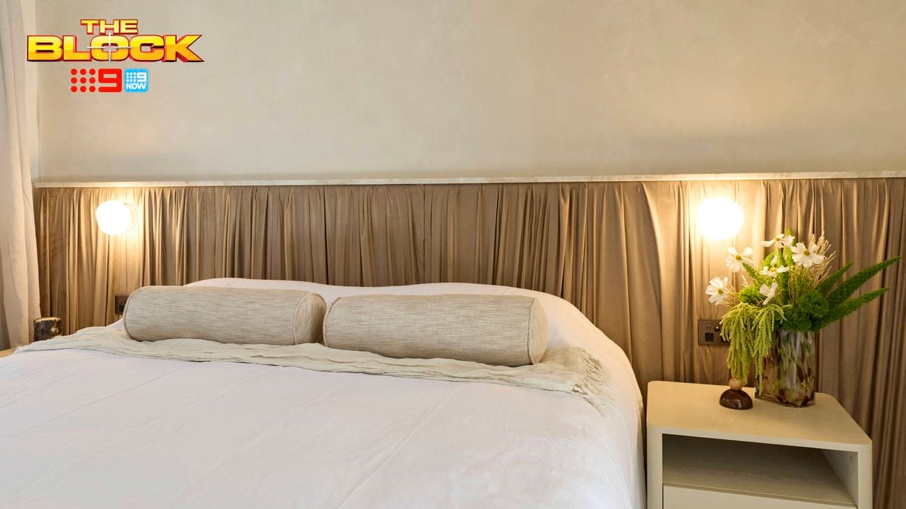 A furnished bedroom with a bed, side table with a vase on it and a bedhead behind it. And The Block logo in the image.