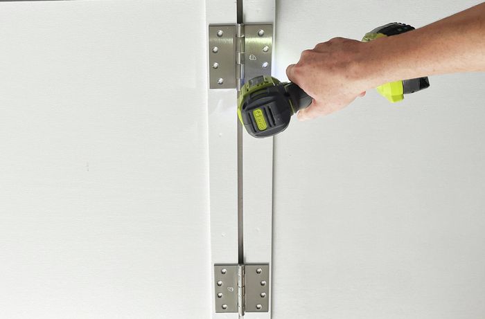 A hand using a drill to attach hinges to two white panels. [[2609] MAK...me sign_V1 | Word]