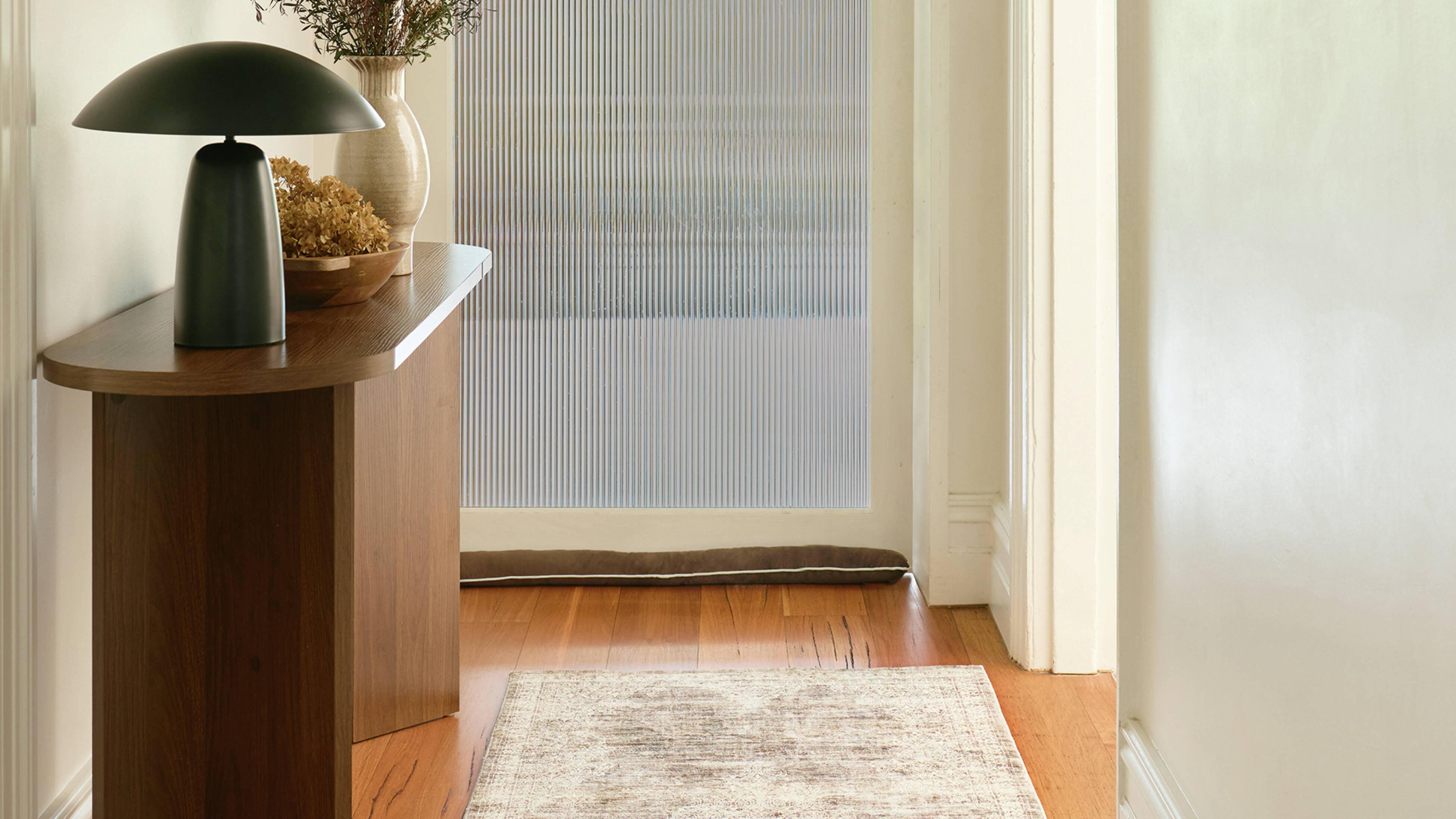 A hallway with a wooden console table, a black mushroom lamp, a vase, a door with reeded glass, a draught stopper, and a runner rug on a wooden floor.