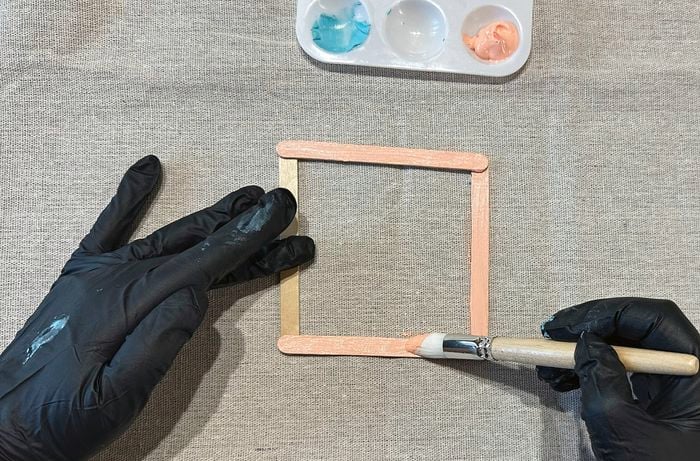 Hands wearing black gloves painting a wooden craft-stick frame with peach-coloured paint, with a paint palette in view.