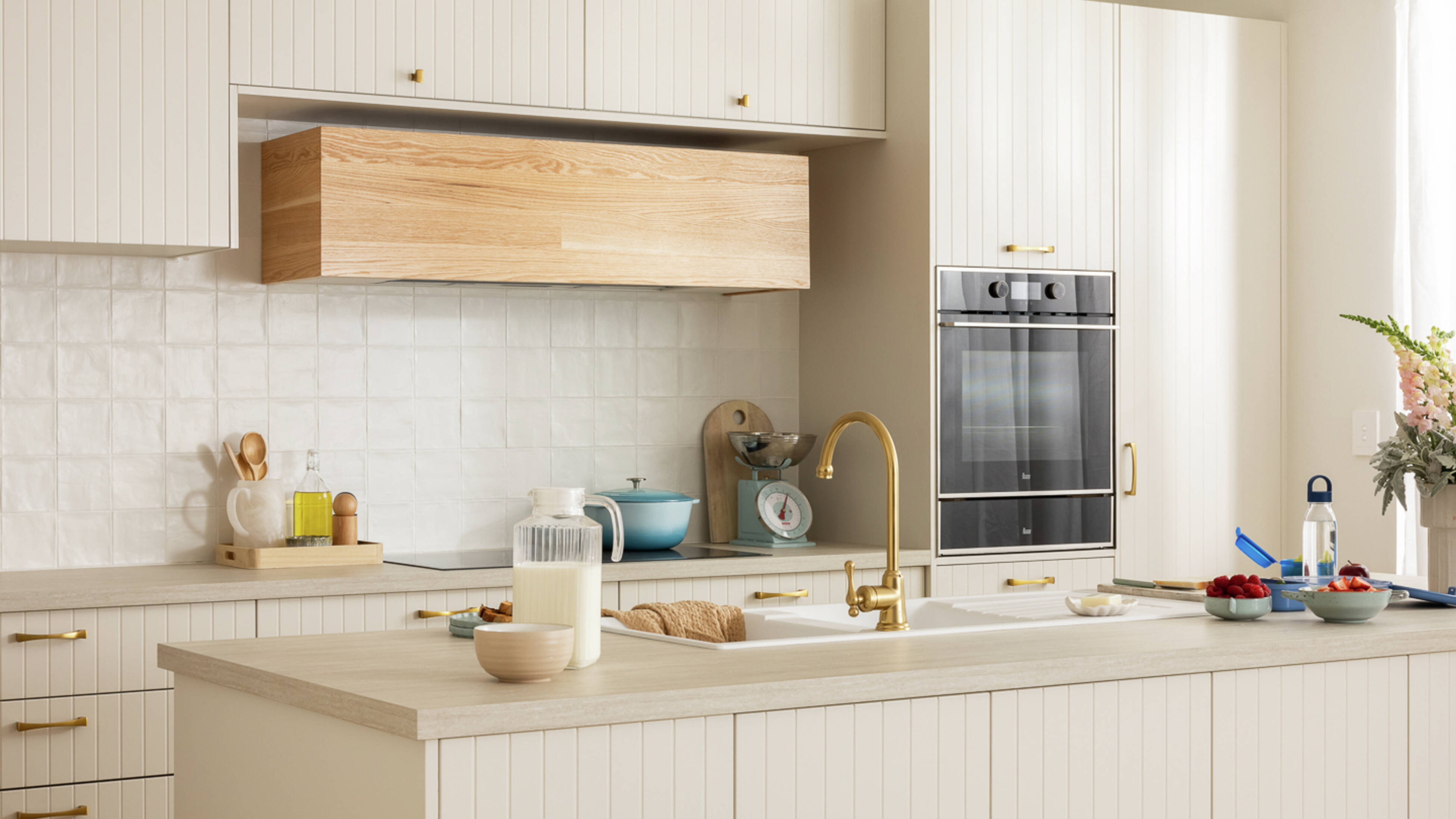Beige modern kitchen with a brown benchtop, gold sink mixer, crockery and a milk jug on the bench.