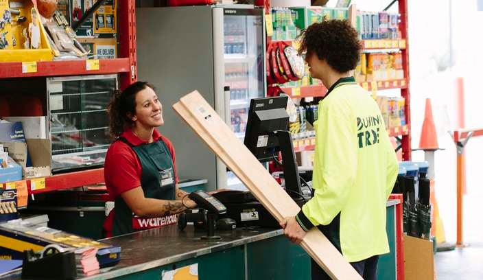A Bunnings Trade specialist carrying lengths of timber talking to a Bunnings team member at a warehouse checkout