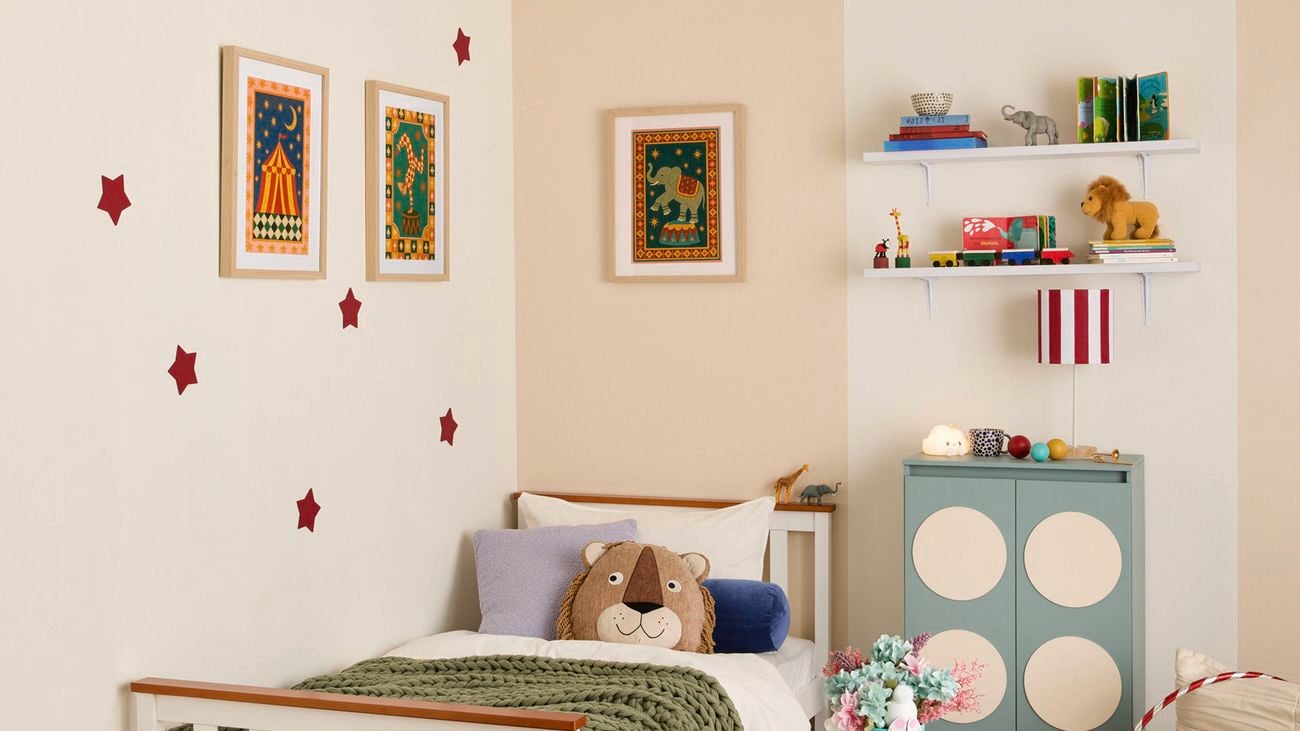 Children’s bedroom with a single bed, framed circus prints and red star wall decals, two white shelves holding books and toys, and a green cabinet with four cream circles topped with a red‑and‑white striped lamp.