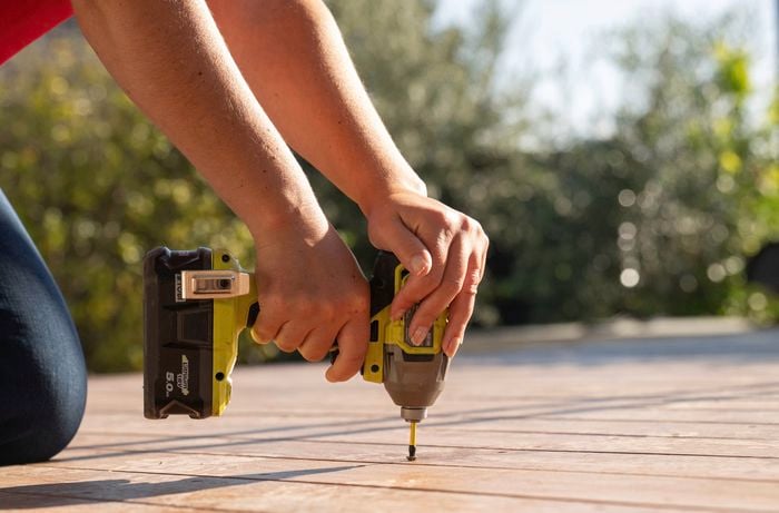 A close-up of a person using a drill to ​tighten deck screws and secure loose boards as part of the preparation process before sanding.​​