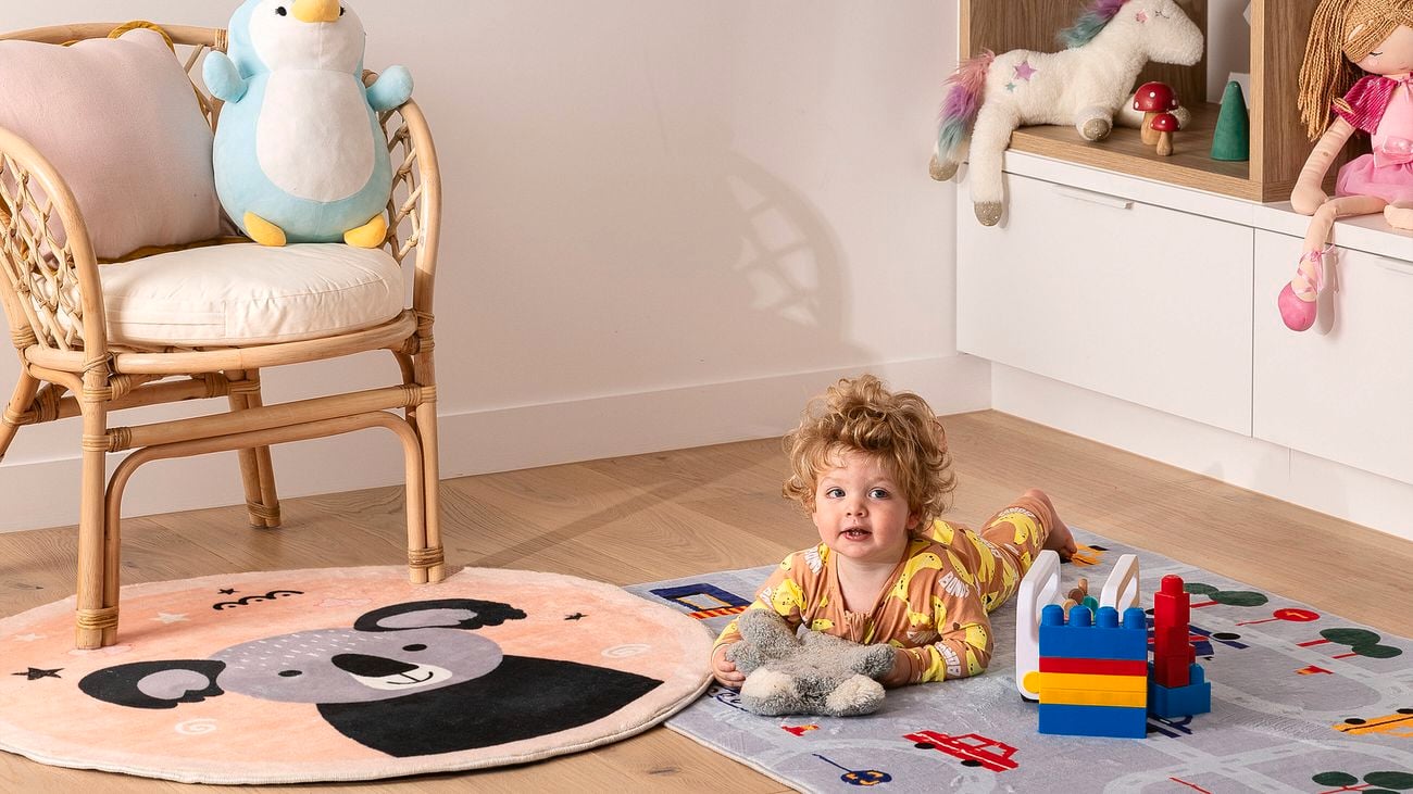 Nursery with wooden floorboards, a woven chair, toys on a shelf. On the floor are two rugs with a baby lying down playing with a grey stuffed animal. One rug has a koala and the other has roads and cars