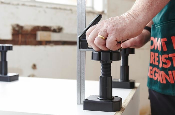 A person using a combination square to set the height of an adjustable foot on a cabinet