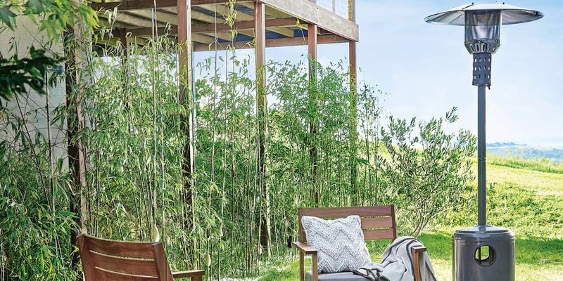 Outdoor living space with patio heater. The area is surrounded by lush green grass and what appears to be bamboo plants in the background. A pair of chairs are positioned by a fire at the centre with a dog in the foreground.